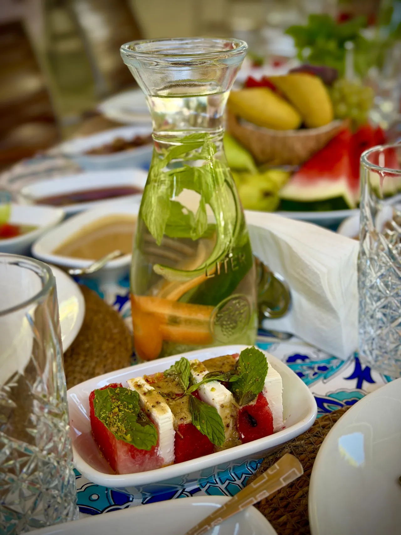 A carafe with citrus fruit and herbs beside a vegetable dish.